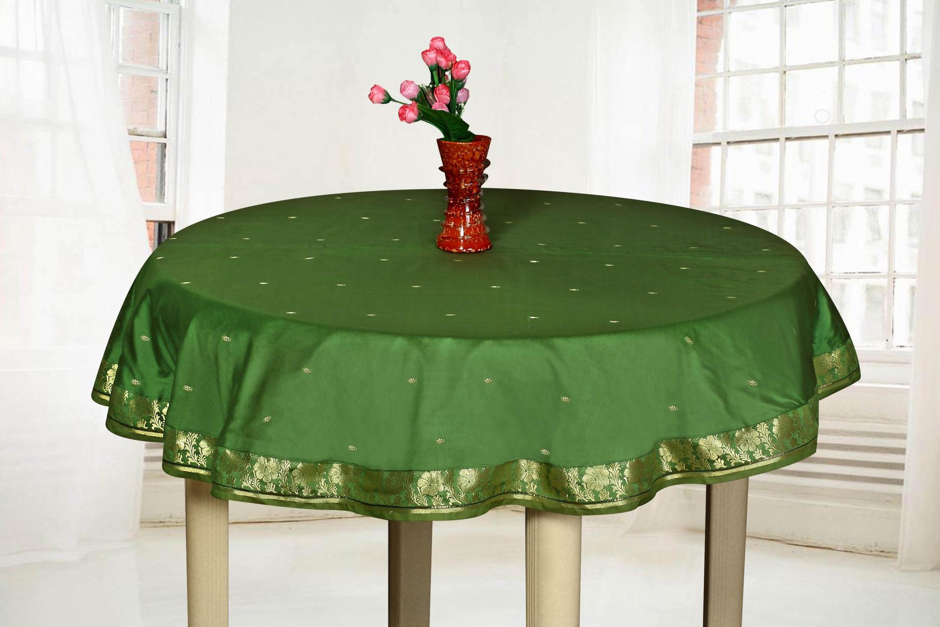Round dark green tablecloth with gold dot pattern and floral border, red glass vase with pink roses, bright airy setting