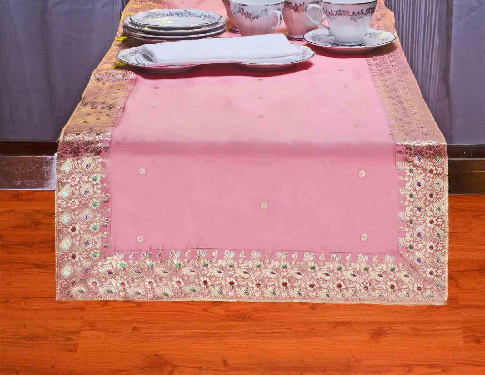 Pink table runner with an intricate gold and multicolor floral border on a wooden floor, set with light-colored dishware, arranged in a bright, airy mood